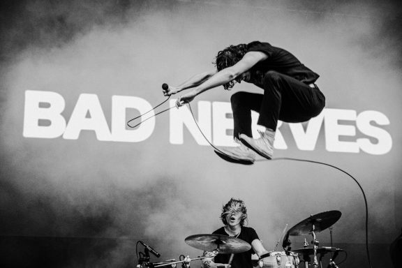 A musician jumps in front of a "BAD NERVES" backdrop, with a drummer visible behind. Bobby Nerves Bad Nerves