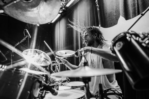 The drummer Ollie Button of UK Foo Fighters passionately playing, with dynamic hair movement and bright stage lights.