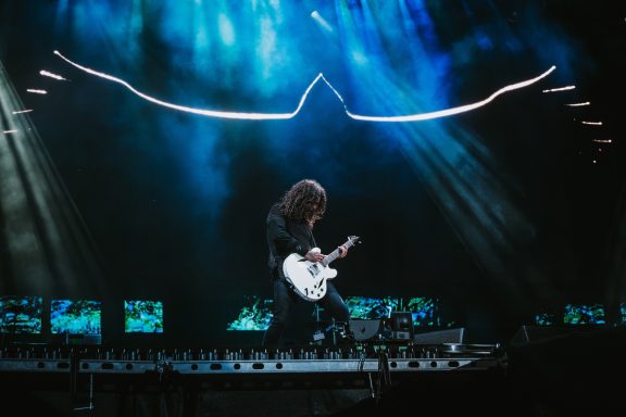 Dave Grohl playing electric guitar on stage with dramatic lighting and a Hawk backdrop design.