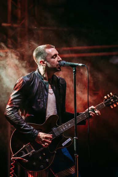 Male musician performing on stage with an electric guitar and microphone, surrounded by smoke.